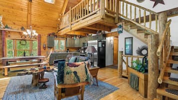 A rustic cabin interior with a wooden staircase, a dining area with benches, a kitchen with modern appliances, and a cozy seating area with a large chair and a blanket.