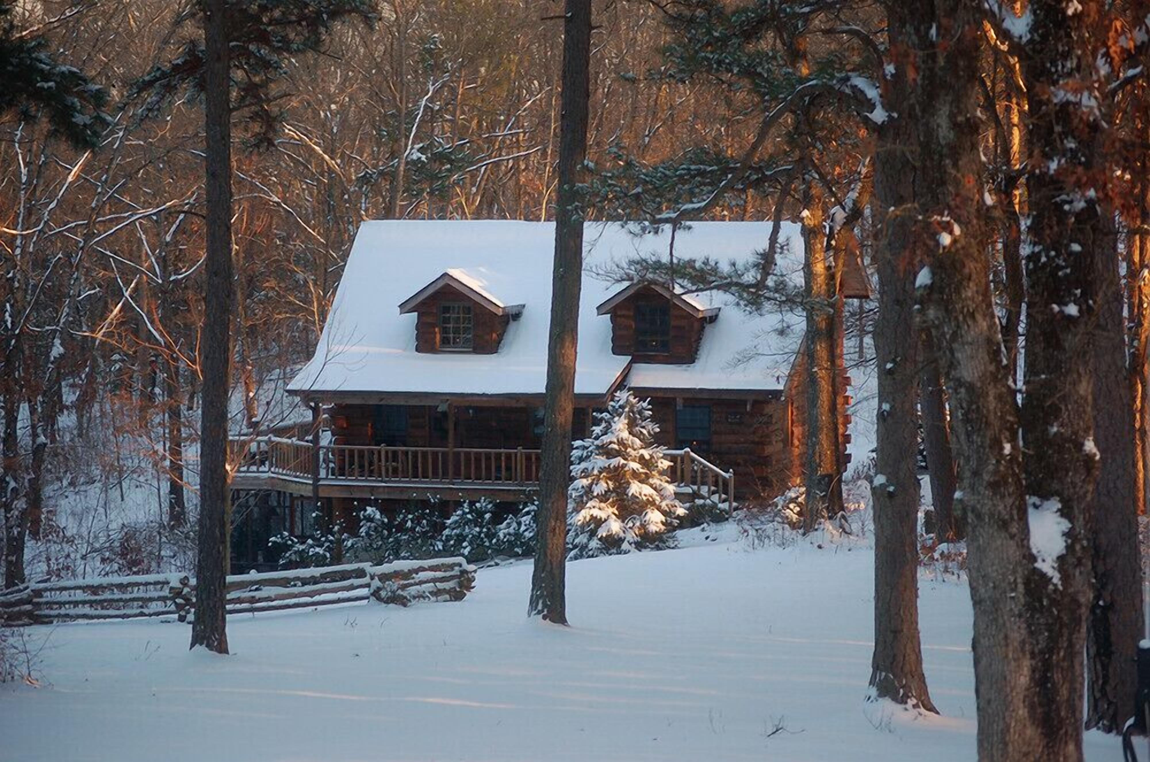 Une cabane en rondins dont le toit est recouvert de neige, entourée d’arbres et dotée d’une terrasse en bois.