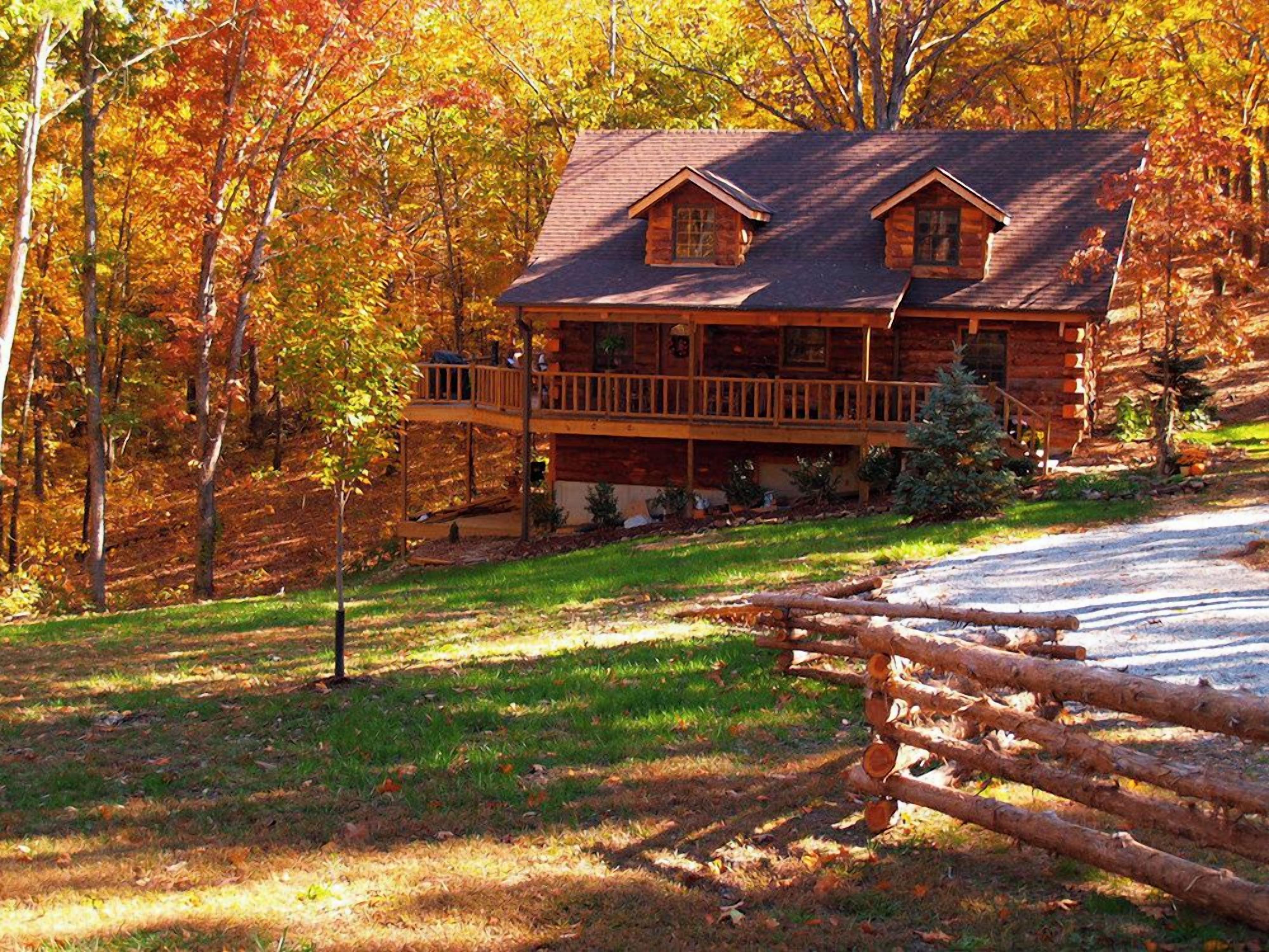 A log cabin with a porch and a deck, surrounded by trees with autumn foliage.