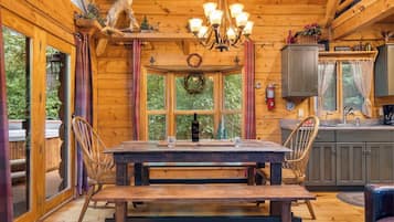 A wooden cabin interior with a dining table, chairs, a chandelier, and a kitchen area.