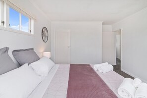 A hotel room with a bed, pillows, a bedside table, a lamp, a door, and a view of the hallway. - Thermal Adventure - Plunge Pool, Sleeps 11 (Rotorua)