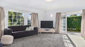 A modern living room with a dark sofa, a flat-screen TV on a wooden TV stand, and large windows with curtains. - Thermal Adventure - Plunge Pool, Sleeps 11 (Rotorua)