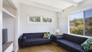 A modern living room with a dark grey sectional sofa, a flat-screen TV on a wooden stand, and a large window offering a view of greenery and buildings. - Arthurs in Mollymook (Mollymook Beach)