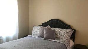 Ein ordentlich bezogenes Bett mit gemusterten Betttextilien, ein dunkles Kopfteil und ein kleiner Nachttisch mit Lampe.