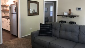 Ein Wohnzimmer mit grauem Sofa, einem schwarz-weißen Kissen, einem Deckenventilator und einer Küchenzeile mit Kühlschrank und Schränken.