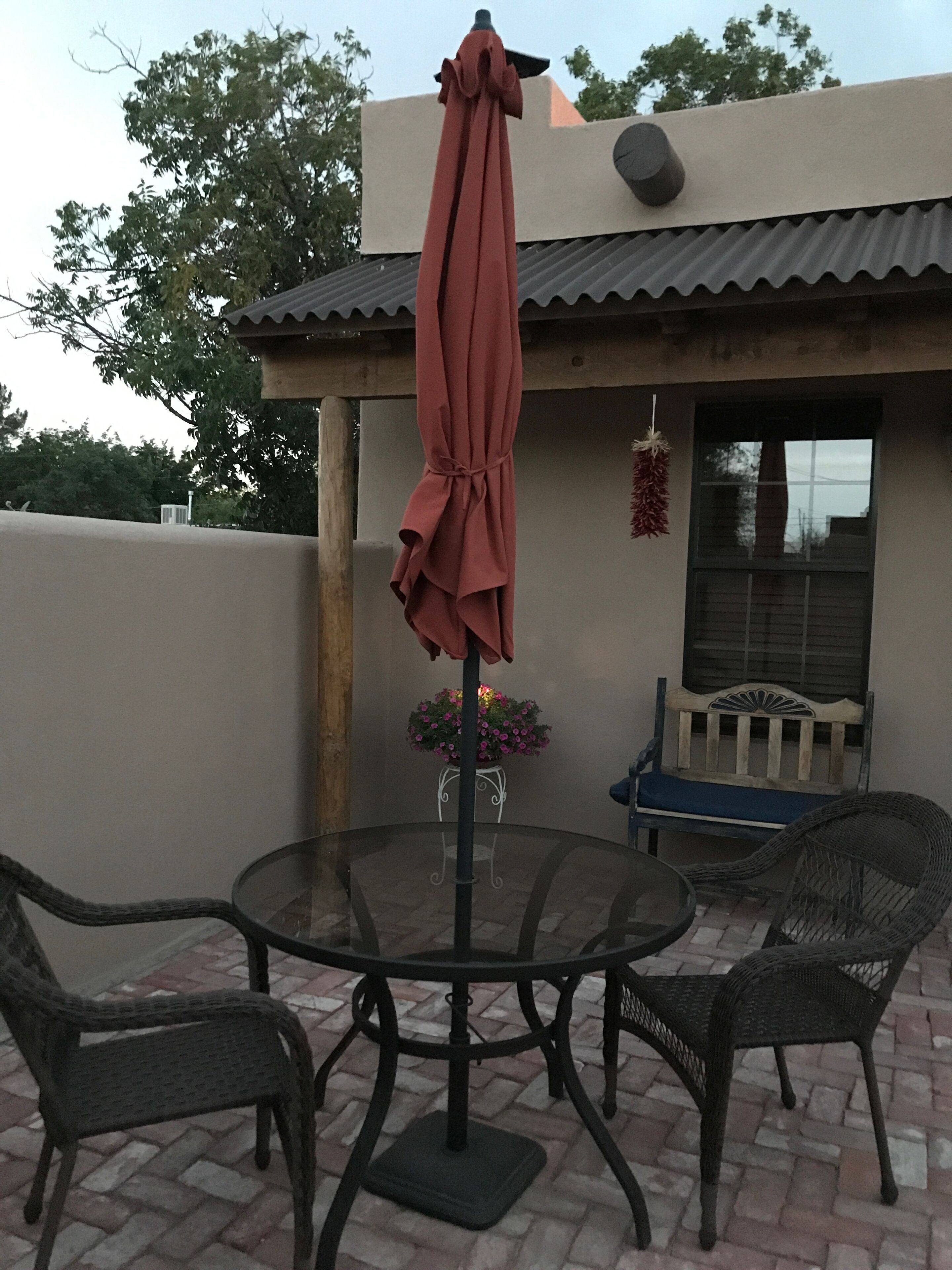 A patio with a round glass table, two wicker chairs, a red umbrella, a small potted plant, and a bench.