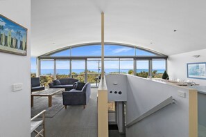 A modern interior with a curved ceiling, a bar area, and a seating arrangement facing a large window with a view of the sea. - Magnum (Lorne)