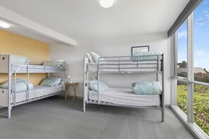 A room with two bunk beds, a small wooden stool, and a large window providing a view of greenery outside.