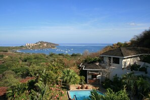 Une maison avec une piscine, entourée d’une végétation tropicale, qui offre une vue sur une zone côtière avec des bateaux et des bâtiments.
