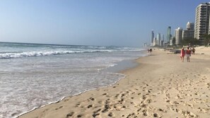 A sandy beach with footprints, a clear sky, and high-rise buildings in the background.