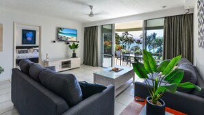A modern living room with a sofa, coffee table, and a potted plant. There is a television on the wall and a balcony with a view of the ocean.