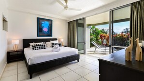 A modern hotel room with a large bed, a ceiling fan, a balcony with a view of tropical plants, and a framed artwork on the wall.