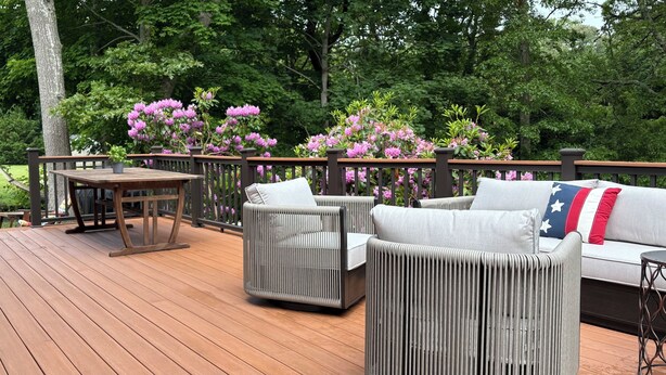 Eine Holzterrasse mit Tisch und Stühlen, eine Sitzecke mit Kissen mit Flaggenmuster und blühenden rosa Blumen.