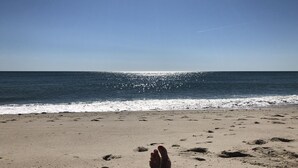 Ein Strand mit Fußabdrücken, die zum Wasser führen, ein klarer Himmel und eine Kondensstrecke eines Flugzeugs.
