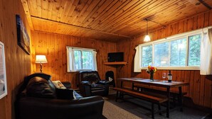 A cozy cabin interior with wooden walls, a leather sofa, a dining table, and a television mounted on the wall. - Amenities & Resort Access Included, Cabin 2 (Phelps)