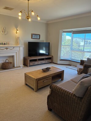 A living room with a television on a wooden stand, a coffee table, a wicker sofa, and a large window. - Ocean Fountain - 100m to the Beach (Moonta Bay)