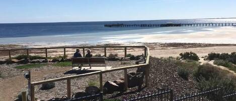 Une plage avec une terrasse en bois et des bancs, une jetée en arrière-plan et un ciel dégagé.