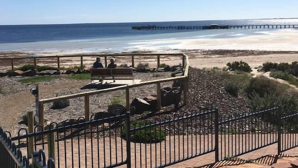 Uma praia com um deck de madeira e bancos, um píer ao fundo e um céu claro.