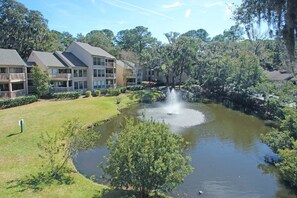 A pond with a fountain, surrounded by lush greenery and a multi-story building with balconies.