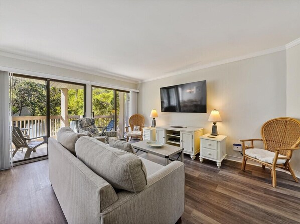 A modern living room with a sofa, a dining table, and chairs. There is a flat-screen TV mounted on the wall and a balcony with a view of trees.