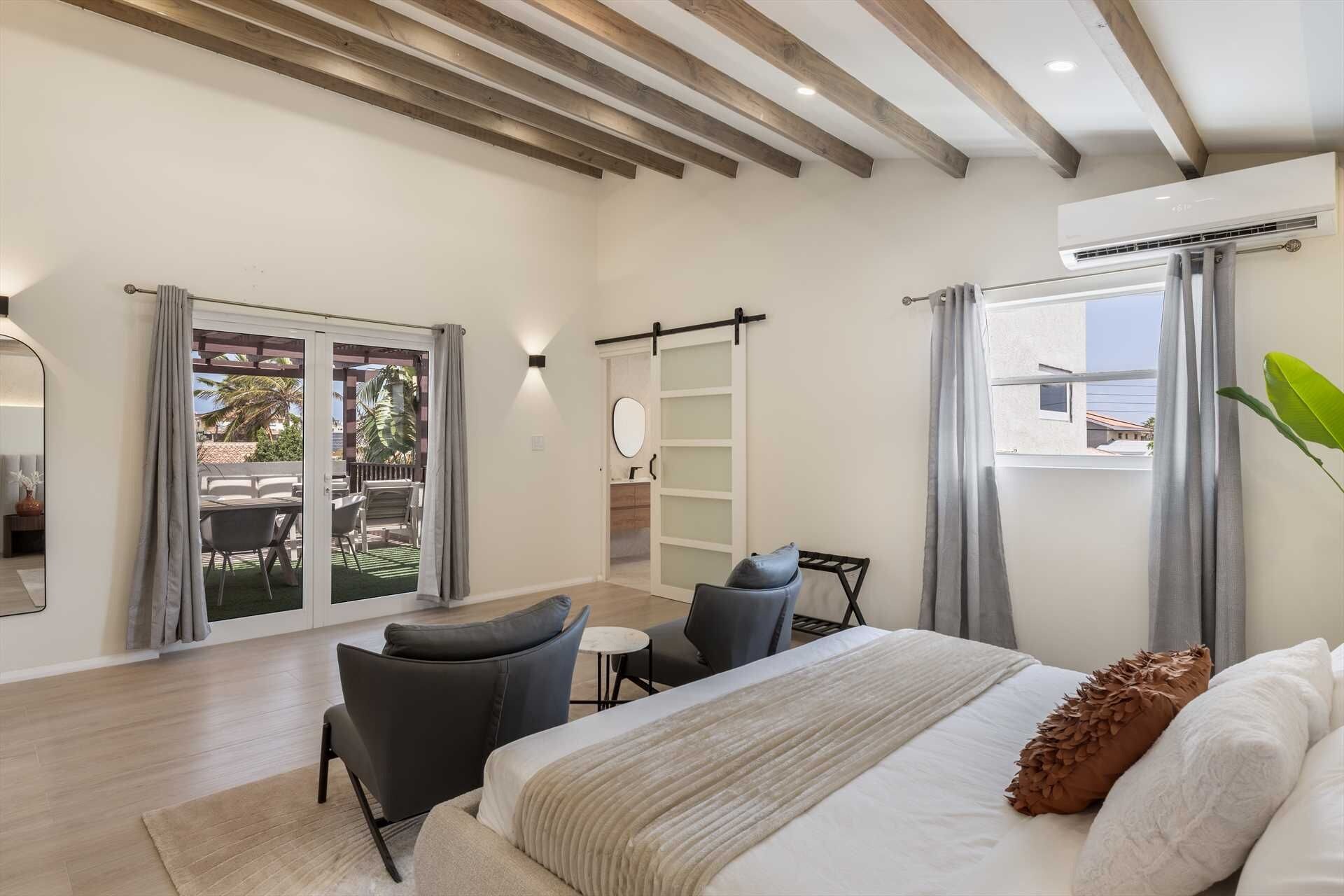 Un dormitorio con cama, dos sillas, una mesita, una puerta corredera a un balcón y un equipo de aire acondicionado.