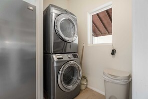 A laundry room with stacked washers and dryers, a wooden broom, a trash can, and a window with a brown roof.