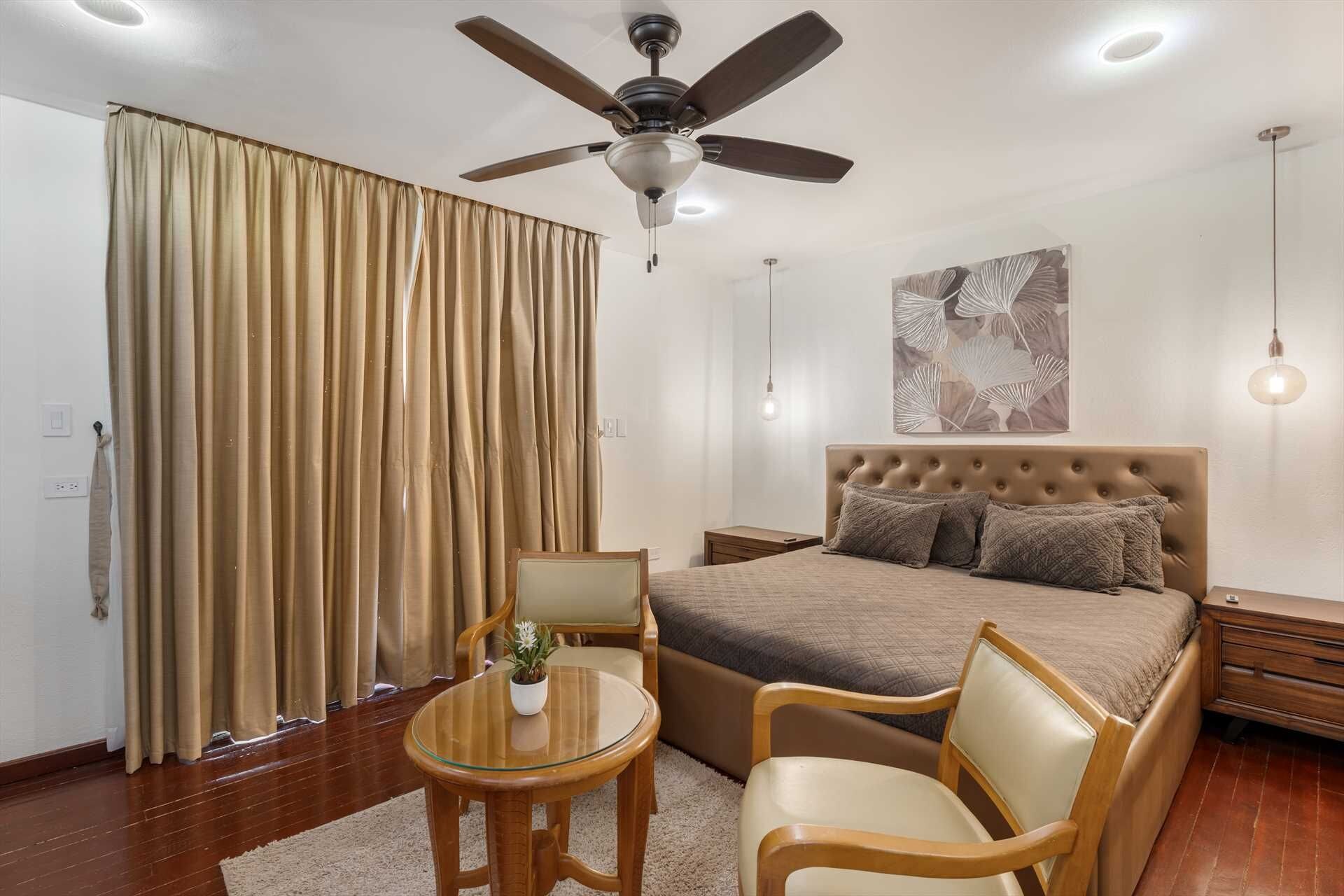 Une chambre à coucher comprenant un lit, deux chaises, une petite table, un ventilateur de plafond et une œuvre d’art murale.