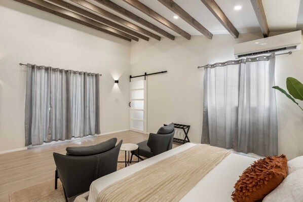 A hotel room with a bed, two chairs, a small table, a plant, and a window with curtains.