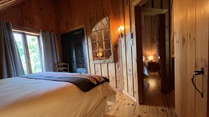 Un dormitorio de cabaña de madera con cama, ventana con cortinas, una puerta y una mesita de noche con lámpara.