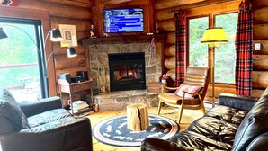 Un acogedor interior de cabaña con chimenea de piedra, sofás de cuero, televisor y cortinas a cuadros.