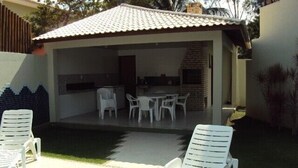 Un patio extérieur couvert, avec des chaises en plastique blanches et une table, une cheminée en briques et un toit en tuiles.