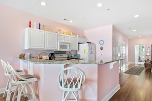 A kitchen with white cabinets, a microwave, a toaster, and a refrigerator. There are two white chairs with patterned cushions. - Seacoast Villa 4 - Unobstructed Ocean View -  Elevator (Folly Beach)