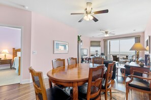 A spacious dining area with a wooden table and chairs, a ceiling fan, and large windows offering a view.