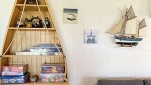 Una habitació amb una unitat de prestatges amb forma de vaixell de fusta plena de joguines i jocs, un sofà gris amb coixins i una petita decoració de veler a la paret.