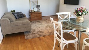 A living room with a grey sofa, a wooden side table with a clock, a glass coffee table with white chairs, and a TV mounted on the wall. - Little Vella, 2 Bedroom 1 bathroom. Secure parking. Free WIFI. Immaculate clean (Ascot Vale)
