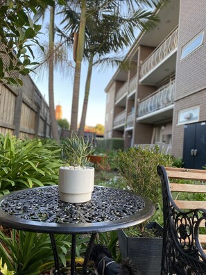Una mesa metálica redonda con una planta en maceta, rodeada de follaje y un banco de madera.