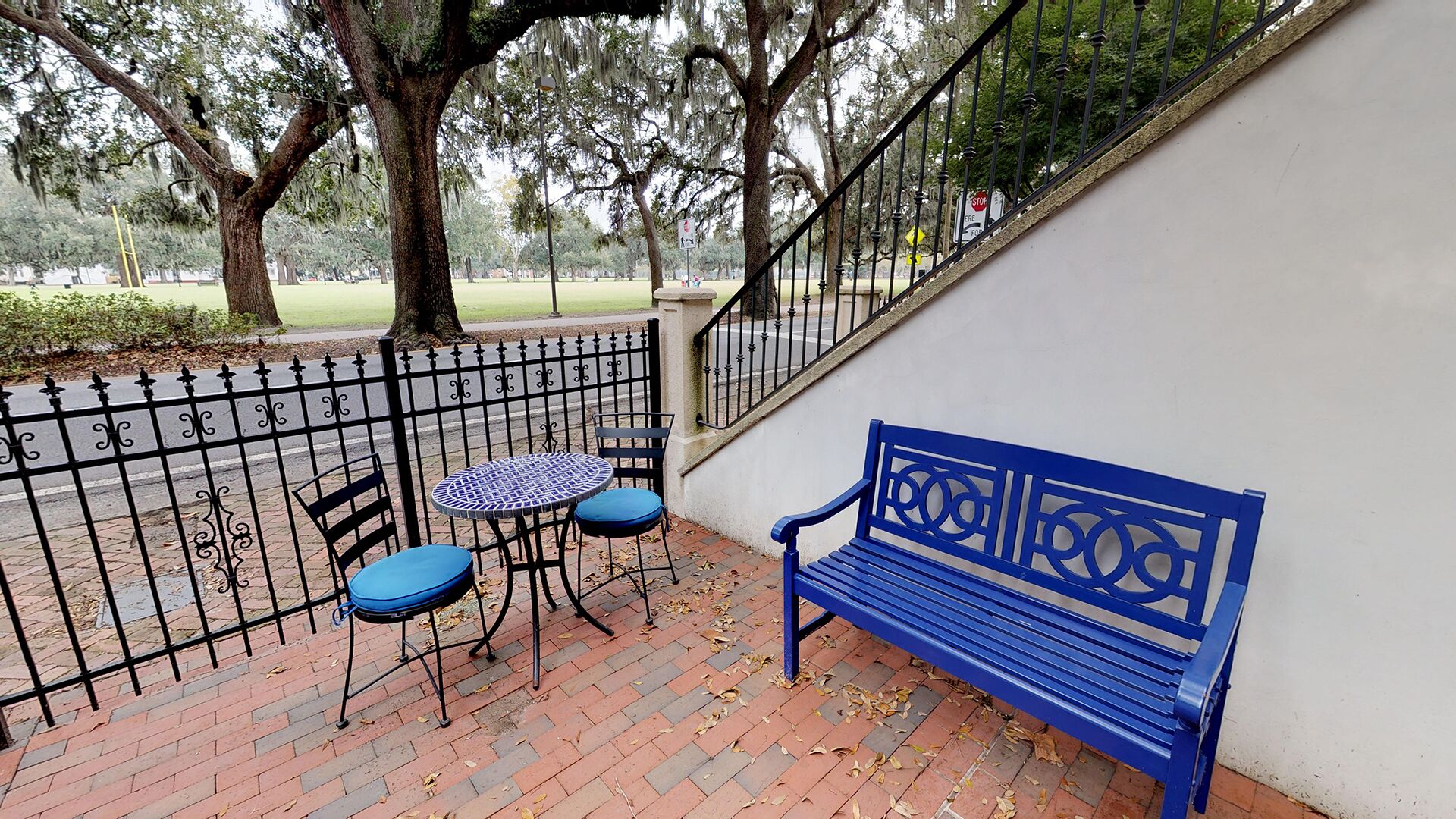 A blue bench and two chairs on a brick patio with a metal fence.