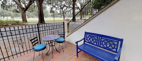 A blue bench and two chairs on a brick patio with a metal fence.