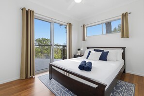 Un dormitorio con cama, cortinas, una mesita de noche, una lámpara y vistas a través de amplios ventanales.