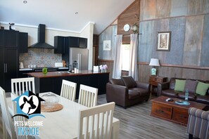 A modern kitchen with dark cabinets, a central island, and a dining area with a table and chairs. - A Beautiful  Property, Walking Distance to the Beach in Esperance WA! (West Beach)