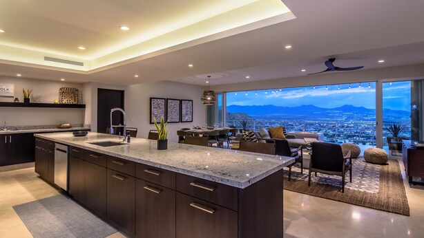 Una cocina moderna con una isla grande, armarios oscuros y vistas a un paisaje urbano a través de amplios ventanales.