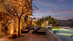 Zona junto a la piscina con sillones de descanso, bañera de hidromasaje y vistas a las montañas al anochecer.