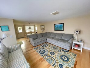 Ein Wohnzimmer mit einem grauen Ecksofa, einem gemusterten Teppich, einem gestreiften Sofa, einem weißen Beistelltisch mit einer Lampe und einem gerahmten Bild an der Wand.
