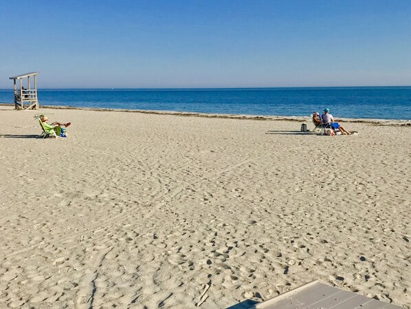 Ein sandiger Strand mit zwei Personen, die auf Liegestühlen sitzen, einem Rettungsschwimmturm und einem wolkenlosen, blauen Himmel.