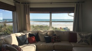 A living room with a beige sofa, various pillows, and a coffee table. A large window offers a view of the beach and ocean. - 4 Bdrm/3 Bath The Tatum House Cottage - Direct Oceanfront (New Smyrna Beach)