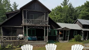 Um braseiro circular com quatro cadeiras Adirondack brancas ao redor, instalado sobre cascalho, com um deck de madeira e um edifício grande de madeira ao fundo.