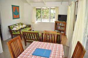 Um quarto com mesa xadrez, cadeiras de madeira, sofá com almofadas estampadas, estante de livros e janela com cortinas.