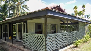 A covered outdoor area with a wooden deck, a door leading inside, and a small porch with a lattice railing. - Rona Kaitangata - across the road from the beach (Muri Beach)