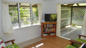 A room with a wooden floor, a television on a stand, a bookshelf, and two green patterned chairs. - Rona Kaitangata - across the road from the beach (Muri Beach)
