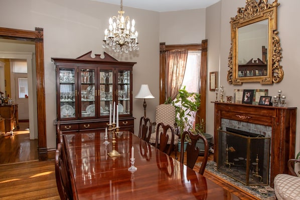 Ein formeller Speisesaal mit Holztisch, Stühlen, einer Chandelüstere, einem Schrank mit Glastüren, einem Kamin und einem großen, verzierten Spiegel.